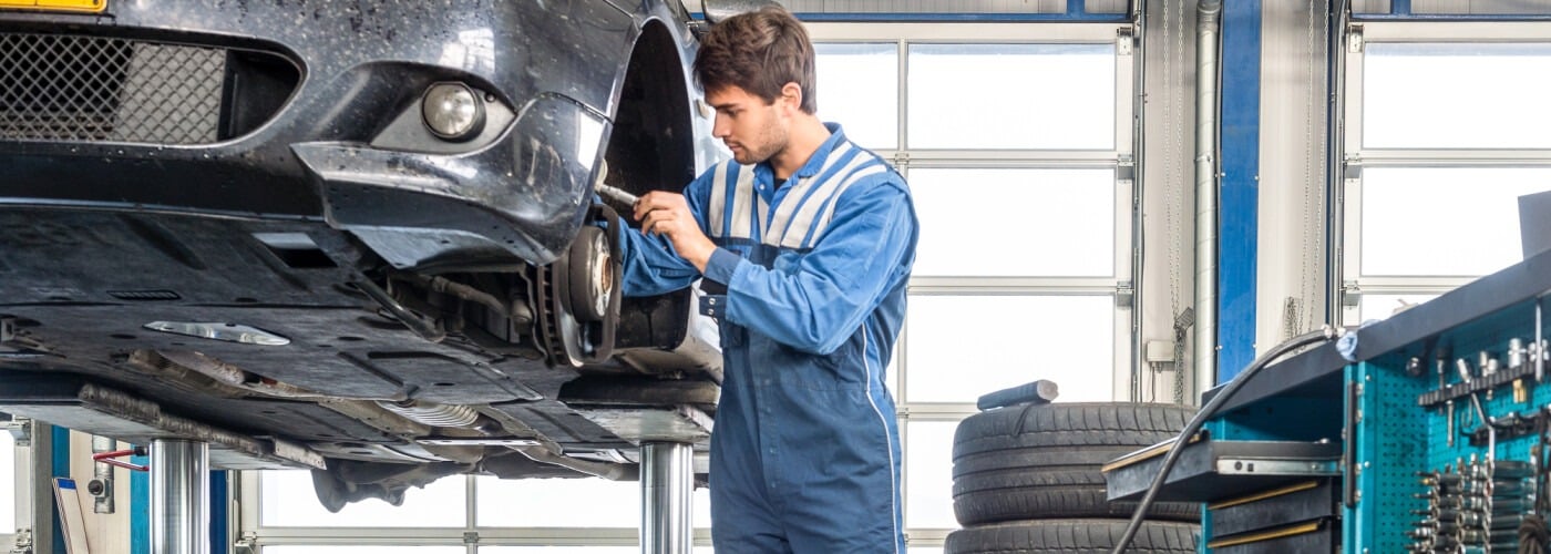 Mechanic in blue coveralls working on car suspension in a garage with tools and tires nearby.