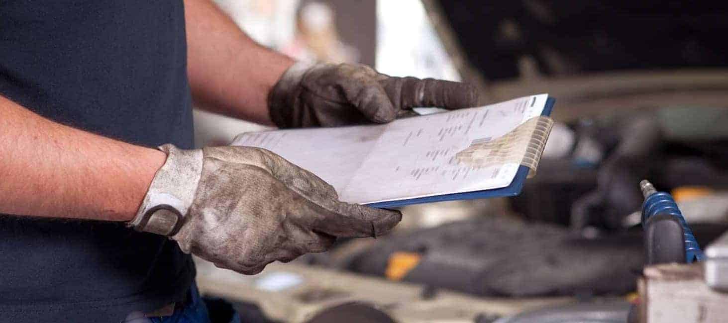 Mechanic with gloves holding a clipboard near an open car hood, performing a vehicle inspection.