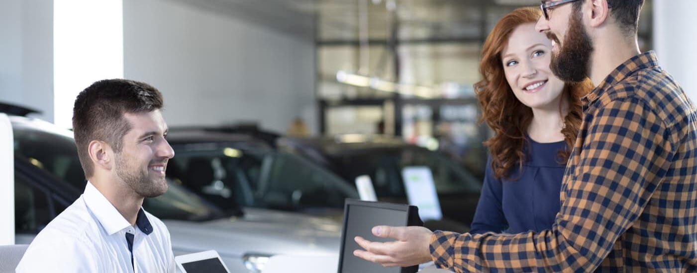 Three people in a car dealership, smiling and conversing, with cars in the background.