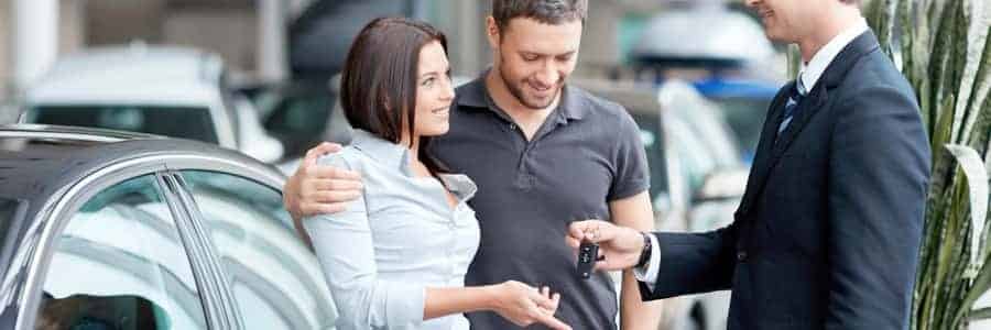 A couple receives car keys from a salesman, standing beside a vehicle in a dealership.
